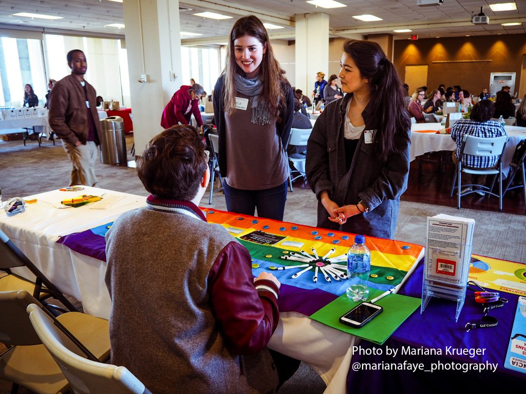 Two attendees stand talking with a person seated at a table with a rainbow-colored tablecloth, with conference attendees seated at tables behind them.